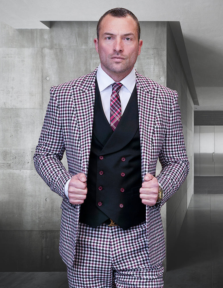 Dressed in the Masimo-Burgundy suit from Statement Clothing, a man stands in a contemporary hallway, confidently adjusting the sleek lapels of his modern-fit houndstooth jacket made from fine Italian wool. He pairs the look with a black vest and a red plaid tie.