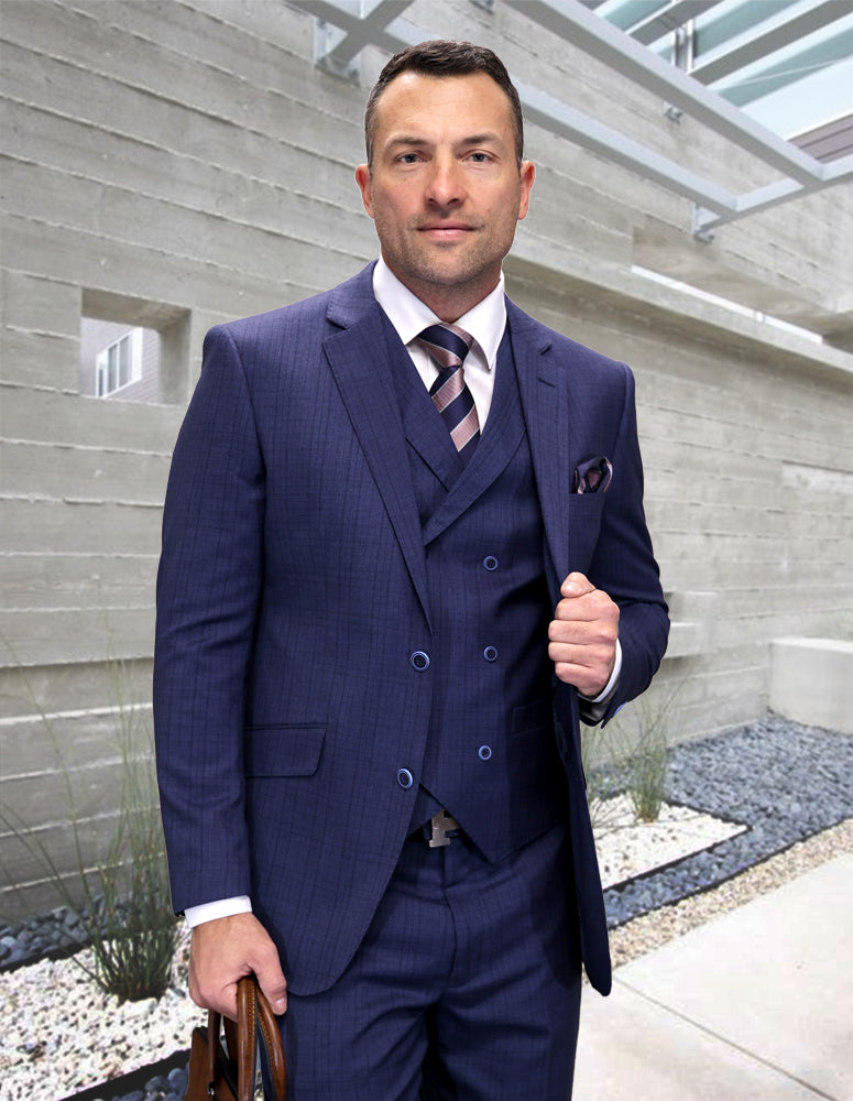 A man in the STATEMENT CLOTHING | MATARI-BLUE Italian wool suit from Statement Clothing, featuring a double-breasted vest, stands outdoors by a modern concrete building holding a brown briefcase and looking at the camera.