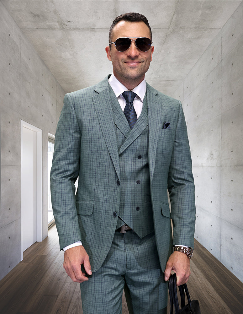 A man, dressed in the STATEMENT CLOTHING | MATARI-SAGE green checked Italian wool suit by Statement Clothing with a double-breasted vest, white shirt, tie, and sunglasses, stands in a modern hallway holding a black bag.