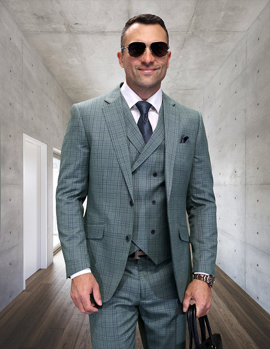 A man, dressed in the STATEMENT CLOTHING | MATARI-SAGE green checked Italian wool suit by Statement Clothing with a double-breasted vest, white shirt, tie, and sunglasses, stands in a modern hallway holding a black bag.