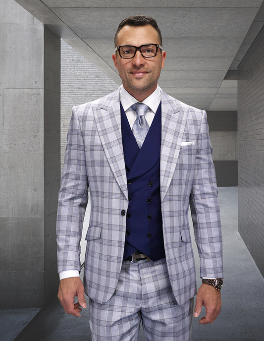 A man wearing Statement Clothing’s MATIAS-GRAY Italian wool plaid suit with modern fit pants, paired with a dark blue vest and patterned tie, stands in a modern concrete hallway.