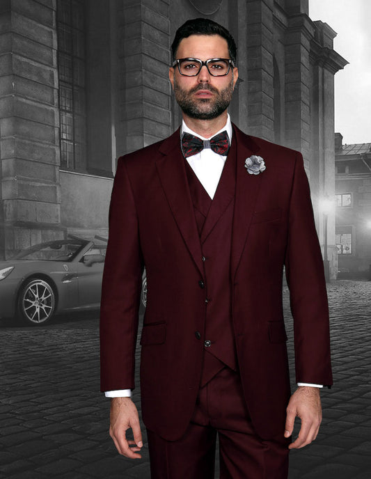 A man in the STATEMENT CLOTHING | MESSINA-BURGUNDY suit by Statement Clothing stands on a cobblestone street, wearing matching vest and bow tie, with pleated pants; a gray building and parked car appear in the background.