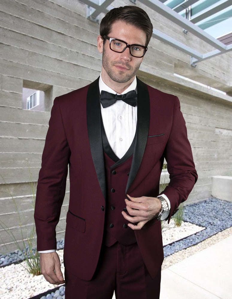 A man in the STATEMENT CLOTHING | MGM-BURGUNDY tailored fit tuxedo with a black lapel by Statement Clothing, paired with a white shirt, black bow tie, and glasses, stands in a modern outdoor setting with concrete and stone landscaping.