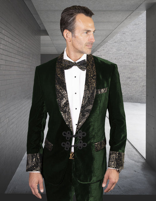 A man in the STATEMENT CLOTHING | MIRAGE-HUNTER tuxedo from Statement Clothing, featuring modern fit dark green velvet with black and gold patterned lapels and cuffs, stands in a sleek gray concrete hallway.