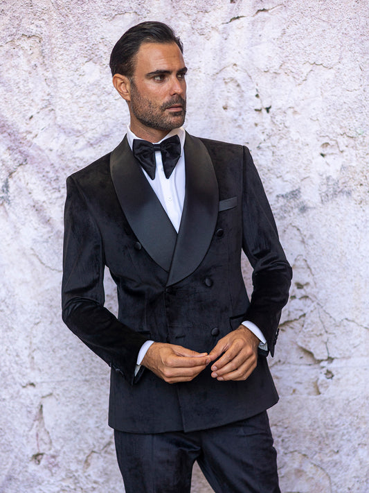 A man in an Insomnia MANZINI MIRAGE 2-Piece Tailored Fit Black Double Breast Velvet Tuxedo stands against a textured wall, looking slightly to the side, his bow tie perfectly complementing his elegant ensemble.