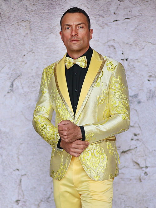 A man stands against a textured wall wearing an Insomnia MANZINI MZS-538 Metallic Glitter Tuxedo Suit Prom Yellow Jacket, featuring a slim fit with floral patterns, paired with a matching bow tie.