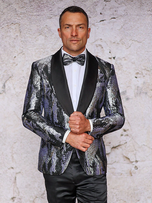A man stands against a light wall, wearing the Manzini MANZINI MZS-552 Men's Textured Metallic Tuxedo Suit Jacket in silver with shawl lapel, paired with a bow tie and holding his jacket with one hand.