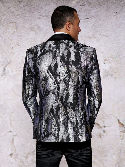 A man stands against a light wall, wearing the Manzini MANZINI MZS-552 Men's Textured Metallic Tuxedo Suit Jacket in silver with shawl lapel, paired with a bow tie and holding his jacket with one hand.