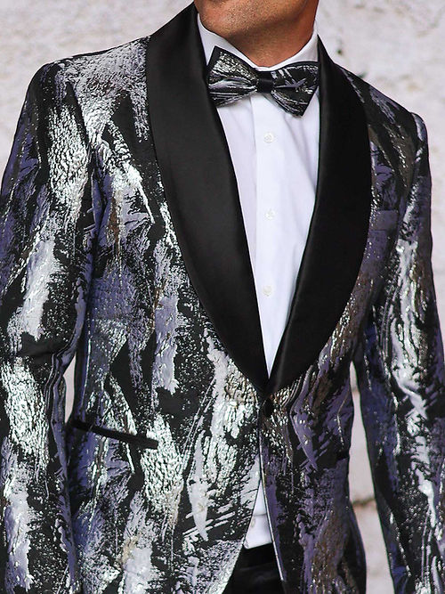 A man stands against a light wall, wearing the Manzini MANZINI MZS-552 Men's Textured Metallic Tuxedo Suit Jacket in silver with shawl lapel, paired with a bow tie and holding his jacket with one hand.