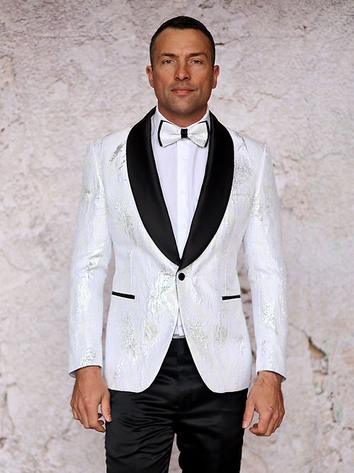 A person wearing an Insomnia MANZINI MZS-552 Textured Metallic Tuxedo Suit Shawl Lapel White Jacket, featuring black lapels and a bow tie in a luxurious poly-rayon blend, stands against a textured light-colored background.
