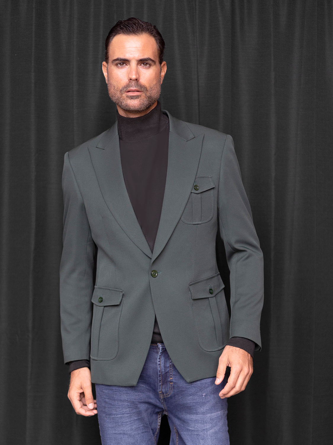 A man wears a Manzini MZS-563 Men's Tailored-Fit 3-Pocket Light Knit Sport Coat Blazer Jacket Hunter over a black turtleneck and blue jeans, standing in front of a dark curtain.