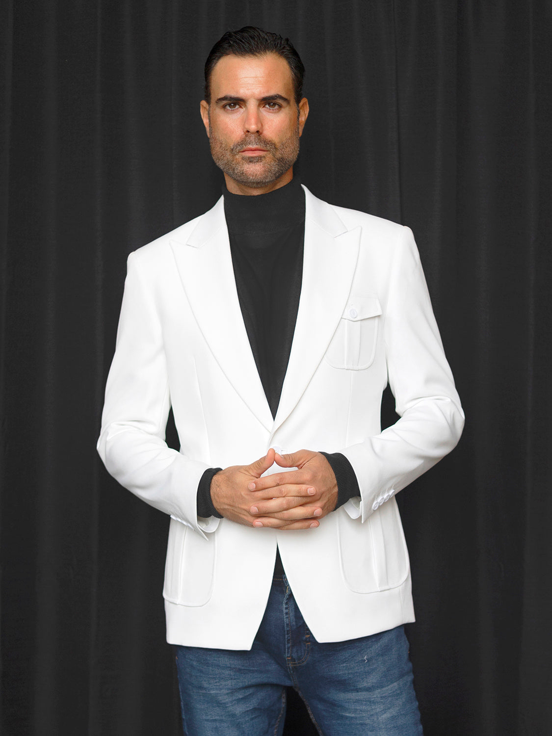 A man stands before a black curtain wearing the Manzini MZS-563 Men's Tailored-Fit 3-Pocket Light Knit Sport Coat Blazer Jacket in white, paired with a black turtleneck and blue jeans, his hands clasped in front of him.