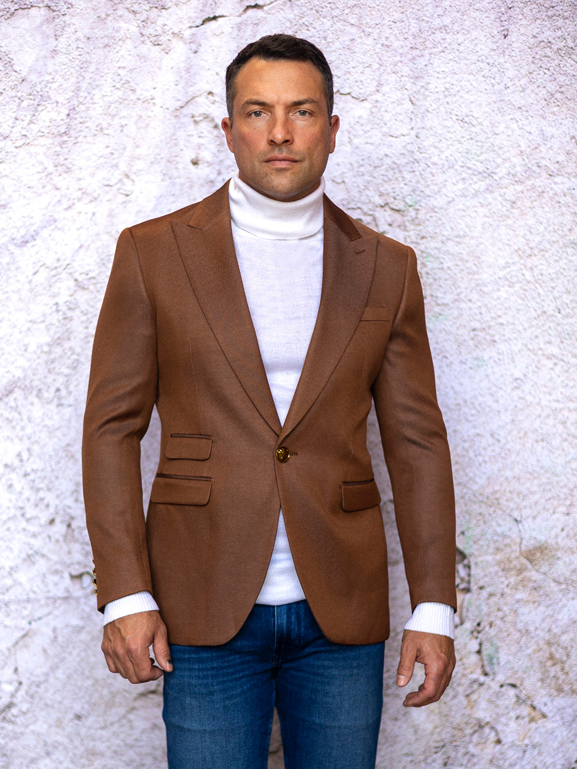 A man wearing a Manzini MZS-564 Men's Tailored-Fit 4-Pocket Light Knit Sport Coat Blazer Jacket in Rust stands before a textured white wall, paired with a white turtleneck sweater and blue jeans, looking at the camera.