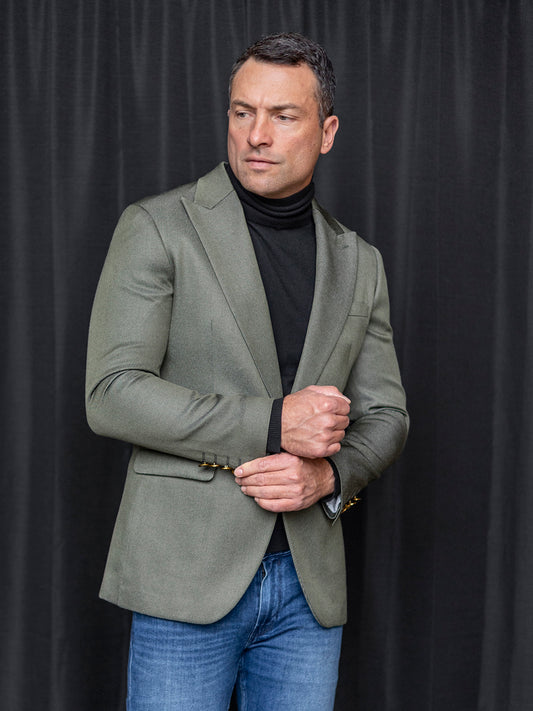A man in a MANZINI MZS-564 4-Pocket Light Knit Hunter Jacket by Insomnia, paired with a black turtleneck and jeans, stands before a dark curtain, adjusting his sleeve.