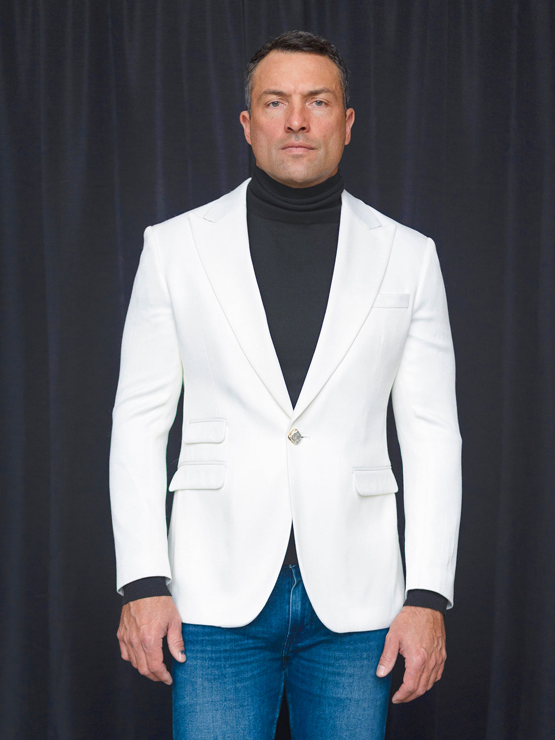 A person stands against a dark curtain, wearing an Insomnia MANZINI MZS-564 4-Pocket Light Knit White Jacket, paired with a black turtleneck and blue jeans.