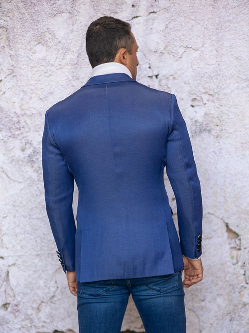 A man wears a Manzini MANZINI MZS-564 Men's Tailored-Fit 4-Pocket Light Knit Sport Coat Blazer Jacket Blue, a white turtleneck sweater, and blue jeans while standing against a textured light-colored wall.