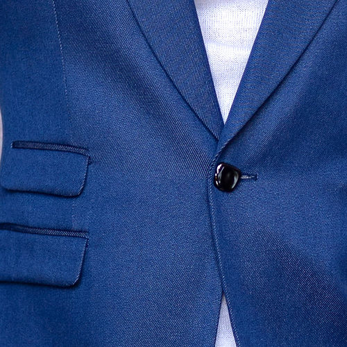 A man wears a Manzini MANZINI MZS-564 Men's Tailored-Fit 4-Pocket Light Knit Sport Coat Blazer Jacket Blue, a white turtleneck sweater, and blue jeans while standing against a textured light-colored wall.