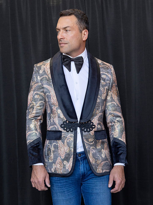 A man wearing a Manzini MZS-566 men's tailored-fit fancy velvet paisley tuxedo dinner jacket in black, paired with a white shirt, black bow tie, and blue jeans, stands in front of a black curtain.
