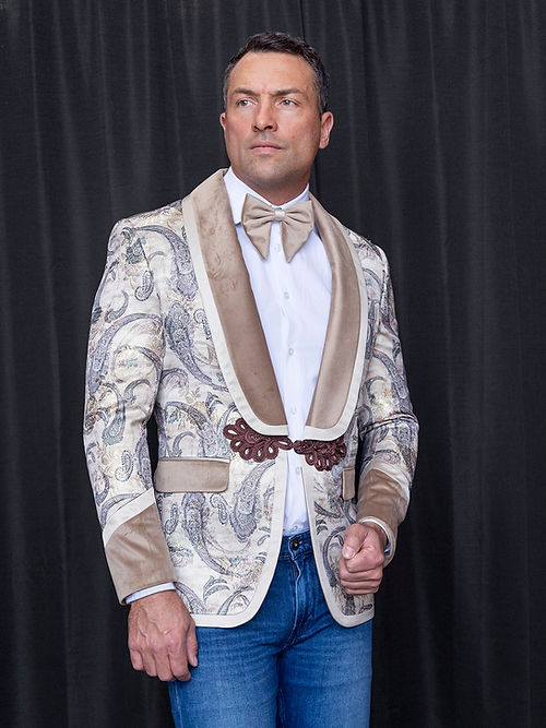 A man wearing the Manzini MANZINI MZS-566 Men's Tailored-Fit Fancy Velvet Paisley Tuxedo Dinner Jacket in brown, paired with beige lapels, a beige bow tie, white shirt, and blue jeans stands before a plain black curtain.
