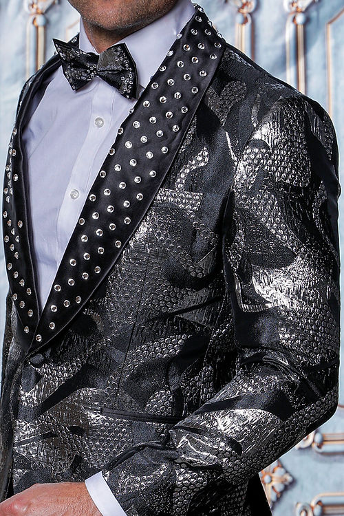 A person is dressed in the Manzini MZS-601 Metallic Pattern Rhinestone Tuxedo Suit featuring a black/silver jacket with rhinestone satin lapels, paired with a black bow tie against a decorative backdrop.
