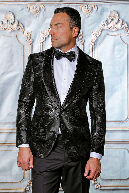 A man wearing a Manzini MZS-601 Black Jacket Tuxedo, featuring a metallic pattern with rhinestones, looks to the side against an ornate backdrop.