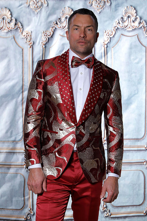 A man in a Manzini MZS-601 Metallic Pattern Rhinestone Tuxedo Suit Burgundy Jacket, featuring a satin lapel and bow tie, stands against a decorative backdrop.