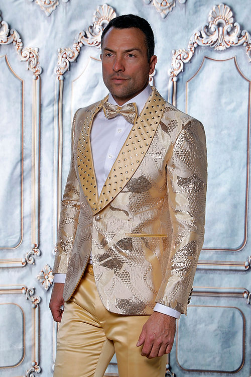 A person in a Manzini MZS-601 Tuxedo Suit featuring a metallic pattern and rhinestone satin lapels stands against an ornate background.