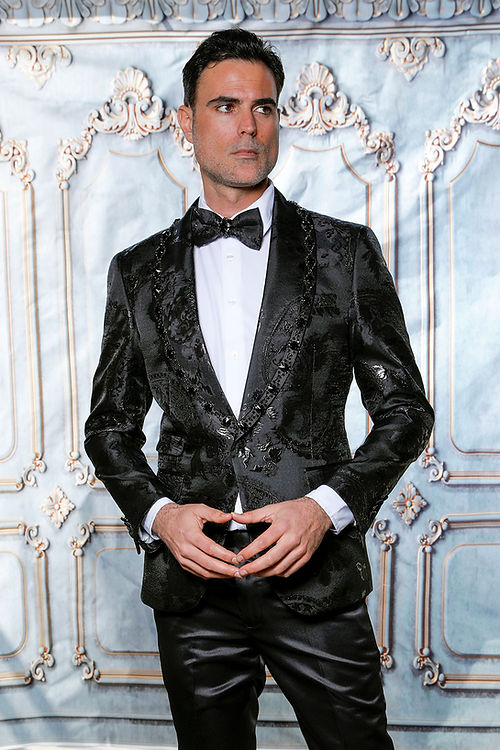 In a Manzini MZS-602 Rhinestone Shiny Metallic Floral Tuxedo Prom Black Jacket, a man exudes elegance with a tailored fit, bow tie, and white shirt, as the shimmering metallic fabric enhances his poised stance with hands together.
