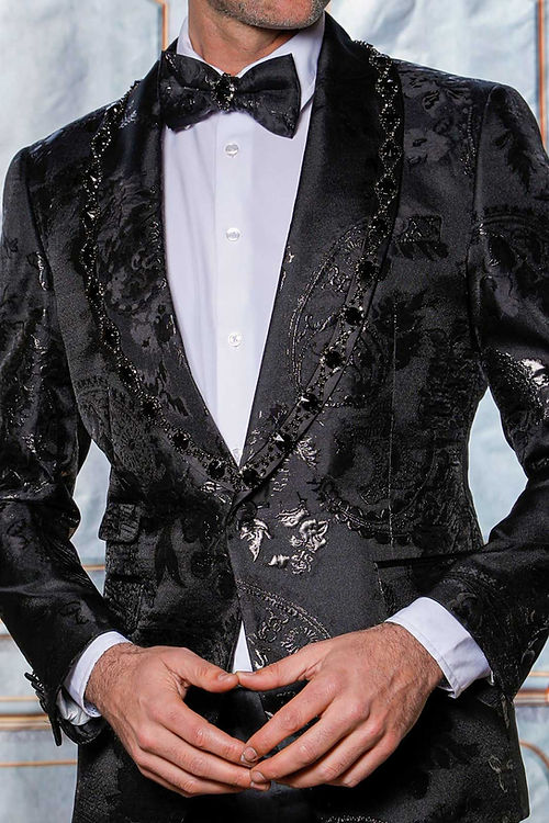In a Manzini MZS-602 Rhinestone Shiny Metallic Floral Tuxedo Prom Black Jacket, a man exudes elegance with a tailored fit, bow tie, and white shirt, as the shimmering metallic fabric enhances his poised stance with hands together.