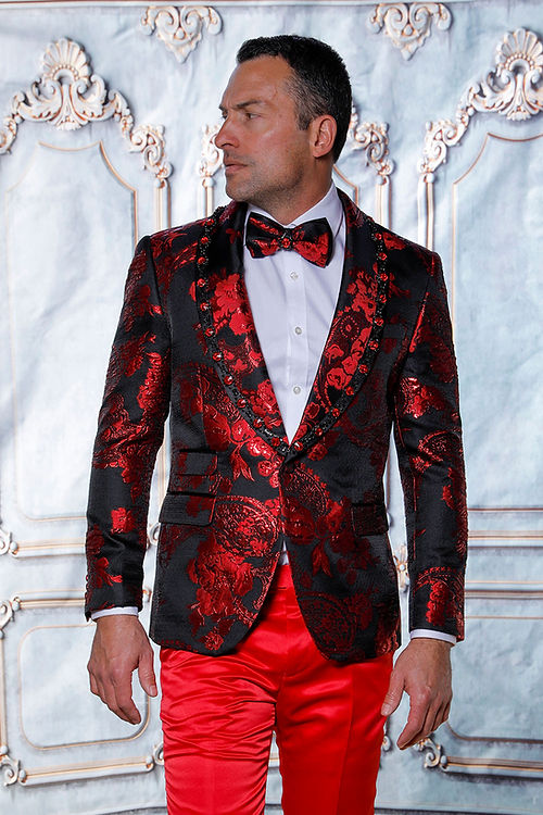 Man wearing a Manzini MZS-602 Rhinestone Shiny Metallic Floral Tuxedo Prom Red Jacket, styled with red pants and a bow tie, stands against an ornate backdrop.