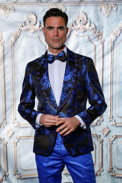 A man wearing a Manzini MZS-602 Rhinestone Shiny Metallic Floral Tuxedo Prom Royal Jacket and bow tie stands against an ornate backdrop where the rhinestone trim subtly catches the light.