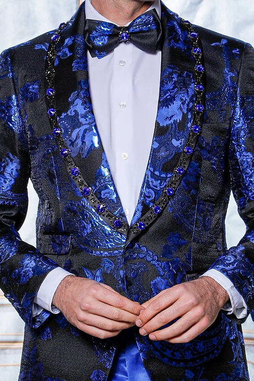A man wearing a Manzini MZS-602 Rhinestone Shiny Metallic Floral Tuxedo Prom Royal Jacket and bow tie stands against an ornate backdrop where the rhinestone trim subtly catches the light.