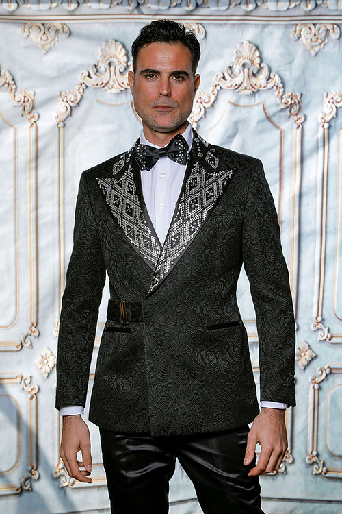 A man in a MANZINI MZS-603 jacket featuring a tailored fit, shiny rhinestones, and a black/silver floral pattern, stands before an ornate light-colored wall.