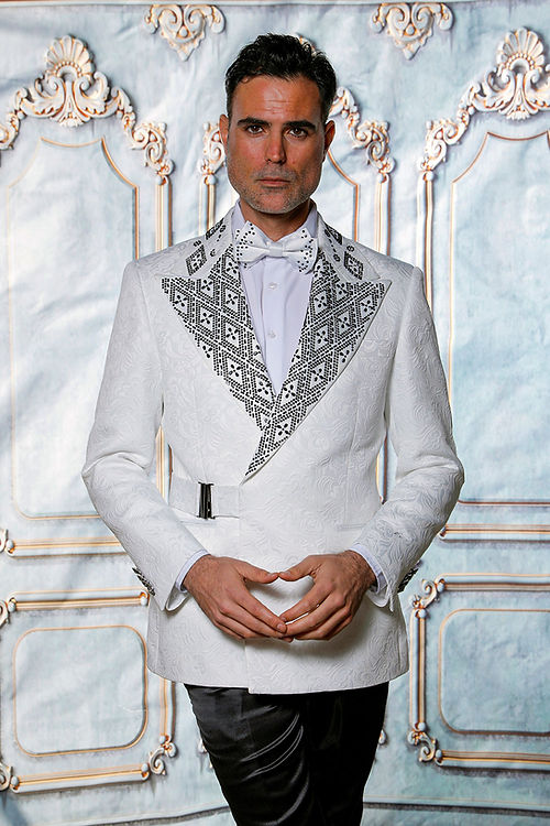 Man in a MANZINI MZS-603 Fancy Shiny Rhinestone Floral Pattern White/Black Jacket by Manzini, standing against a decorative wall.