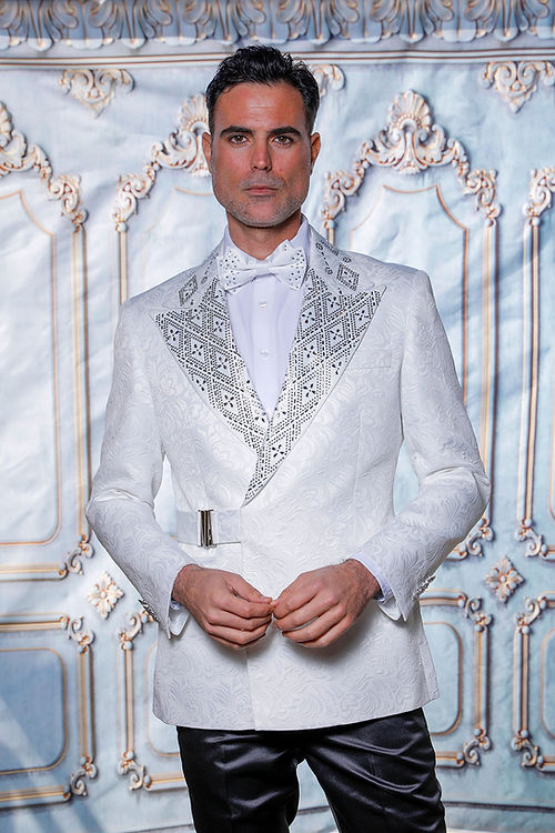 A man wears the Manzini JACKET 37 CLEARANCE—a double-breasted, patterned white tuxedo jacket with metallic lapels and a bow tie—standing before an ornate, light blue and gold decorative backdrop. The jacket’s subtle floral textured pattern enhances the depth and sophistication of his ensemble.