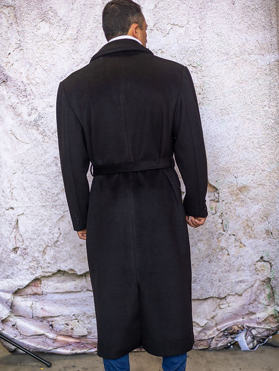 A man stands against a textured wall wearing an Insomnia MANZINI MZW-500 black full-length soft fabric trench coat and a white turtleneck.