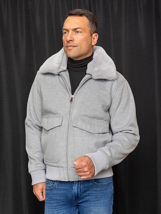 A person stands against a black curtain, wearing the Insomnia MANZINI MZW-501 Grey Bomber Jacket Coat with a removable faux fur collar, paired perfectly with blue jeans for a stylish yet versatile look.
