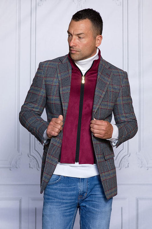 A man stands in front of a white paneled wall, dressed in an Insomnia MANZINI MZW-535 Charcoal Faux Wool Plaid Blazer Coat with Liner, maroon zip-up jacket, white turtleneck, and blue jeans.