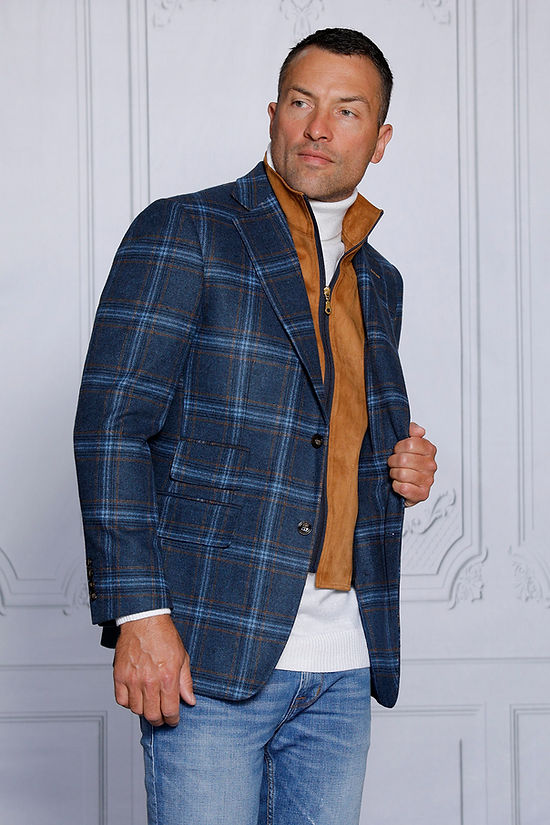 A man in an Insomnia MANZINI MZW-535 indigo plaid blazer coat with a tailored fit, white turtleneck, brown vest, and jeans stands against a light-patterned wall, looking to the side.