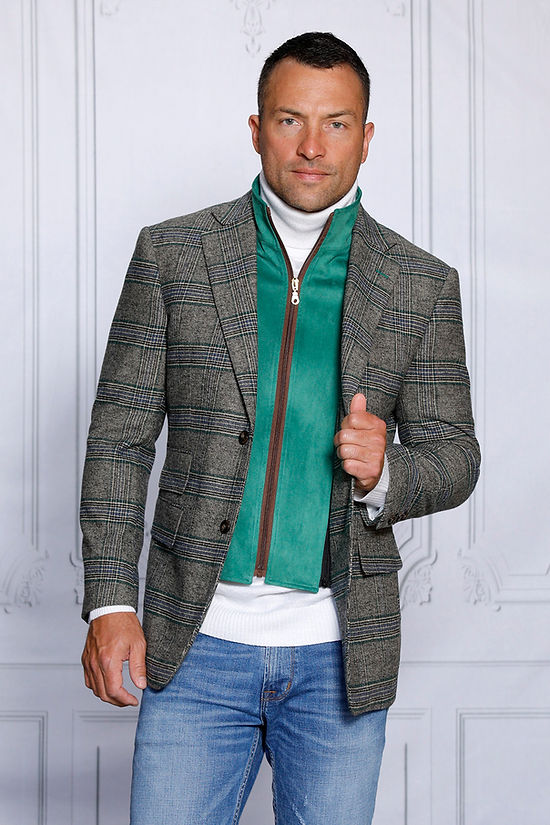 A man confidently stands against a white-paneled wall, wearing an Insomnia tailored fit MANZINI MZW-535 Kelly Green Faux Wool Plaid Blazer Coat and a white turtleneck, complemented by jeans.