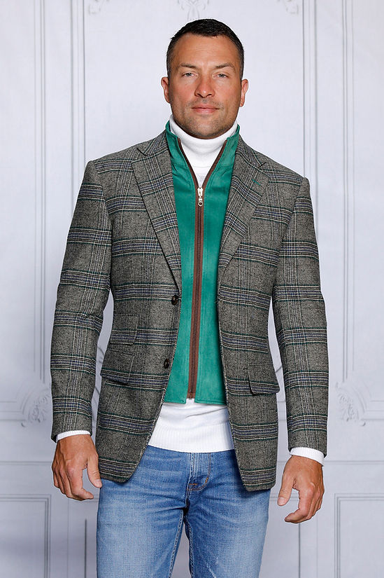 A man confidently stands against a white-paneled wall, wearing an Insomnia tailored fit MANZINI MZW-535 Kelly Green Faux Wool Plaid Blazer Coat and a white turtleneck, complemented by jeans.