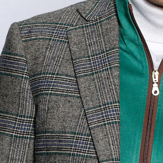 A man confidently stands against a white-paneled wall, wearing an Insomnia tailored fit MANZINI MZW-535 Kelly Green Faux Wool Plaid Blazer Coat and a white turtleneck, complemented by jeans.