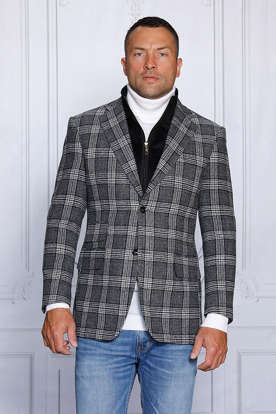 A man stands in front of a gray paneled wall wearing an Insomnia MANZINI MZW-536 black tailored fit faux wool plaid blazer with a zip-out liner over a white turtleneck and zippered vest, paired with blue jeans.
