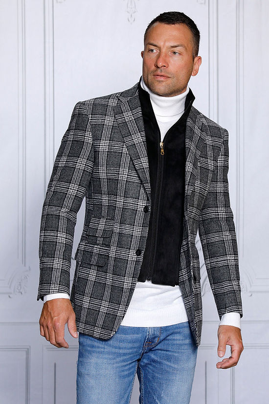 A man stands in front of a gray paneled wall wearing an Insomnia MANZINI MZW-536 black tailored fit faux wool plaid blazer with a zip-out liner over a white turtleneck and zippered vest, paired with blue jeans.