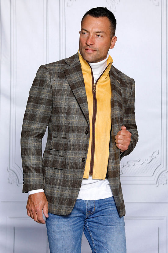 A man wears the Insomnia MANZINI MZW-536 Taupe Tailored Fit Faux Wool Plaid Blazer with a removable zip-out liner over a white turtleneck and yellow vest, confidently paired with blue jeans, set against a textured white wall.