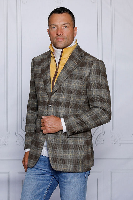 A man wears the Insomnia MANZINI MZW-536 Taupe Tailored Fit Faux Wool Plaid Blazer with a removable zip-out liner over a white turtleneck and yellow vest, confidently paired with blue jeans, set against a textured white wall.