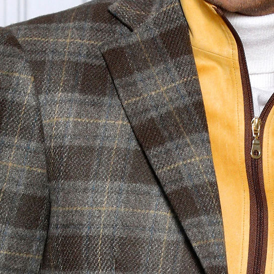 A man wears the Insomnia MANZINI MZW-536 Taupe Tailored Fit Faux Wool Plaid Blazer with a removable zip-out liner over a white turtleneck and yellow vest, confidently paired with blue jeans, set against a textured white wall.