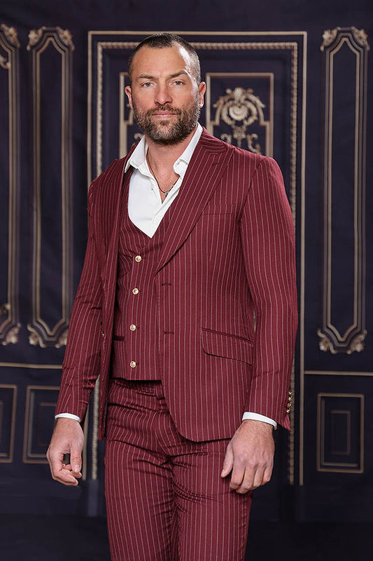 A bearded man wears the Insomnia MUCCLY 3PC Modern Fit Burgundy Thin Pinstripe Suit with a white shirt, standing before an ornate dark backdrop with gold accents and a subtle floral woven pattern.