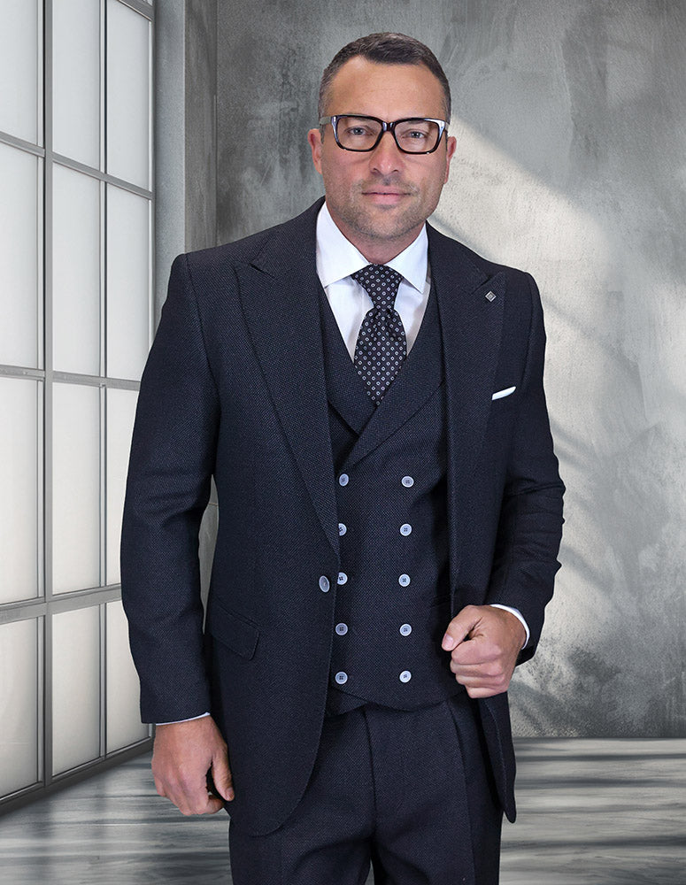 A man wears the STATEMENT CLOTHING | NEIVE-BLACK suit by Statement Clothing, paired with a white shirt and patterned tie, standing indoors against a modern gray textured wall and large window.