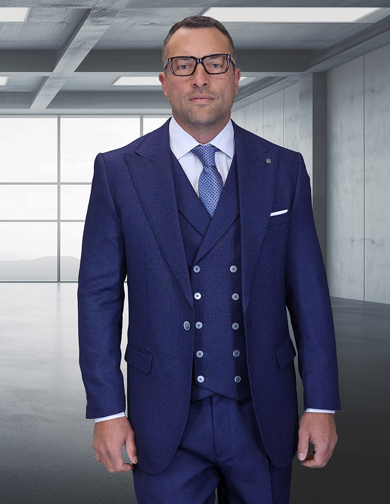 A man in a STATEMENT CLOTHING | NEIVE-SAPPHIRE suit by Statement Clothing, with a light blue shirt, patterned tie, and glasses, stands in a modern empty room with large windows and concrete walls.